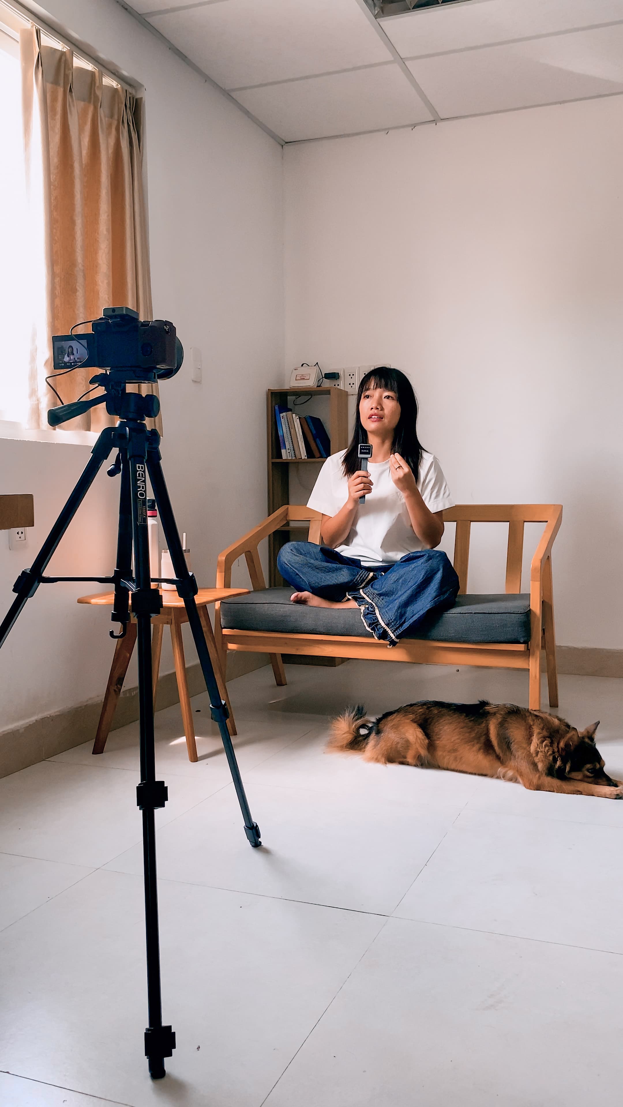 Xuân ngồi trên ghế gỗ tại nhà, nói trước máy quay đặt trên tripod bên cửa sổ, một chú chó nằm nghỉ trên sàn